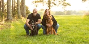 Man and woman crouching down holding dogs in a sunny forest area